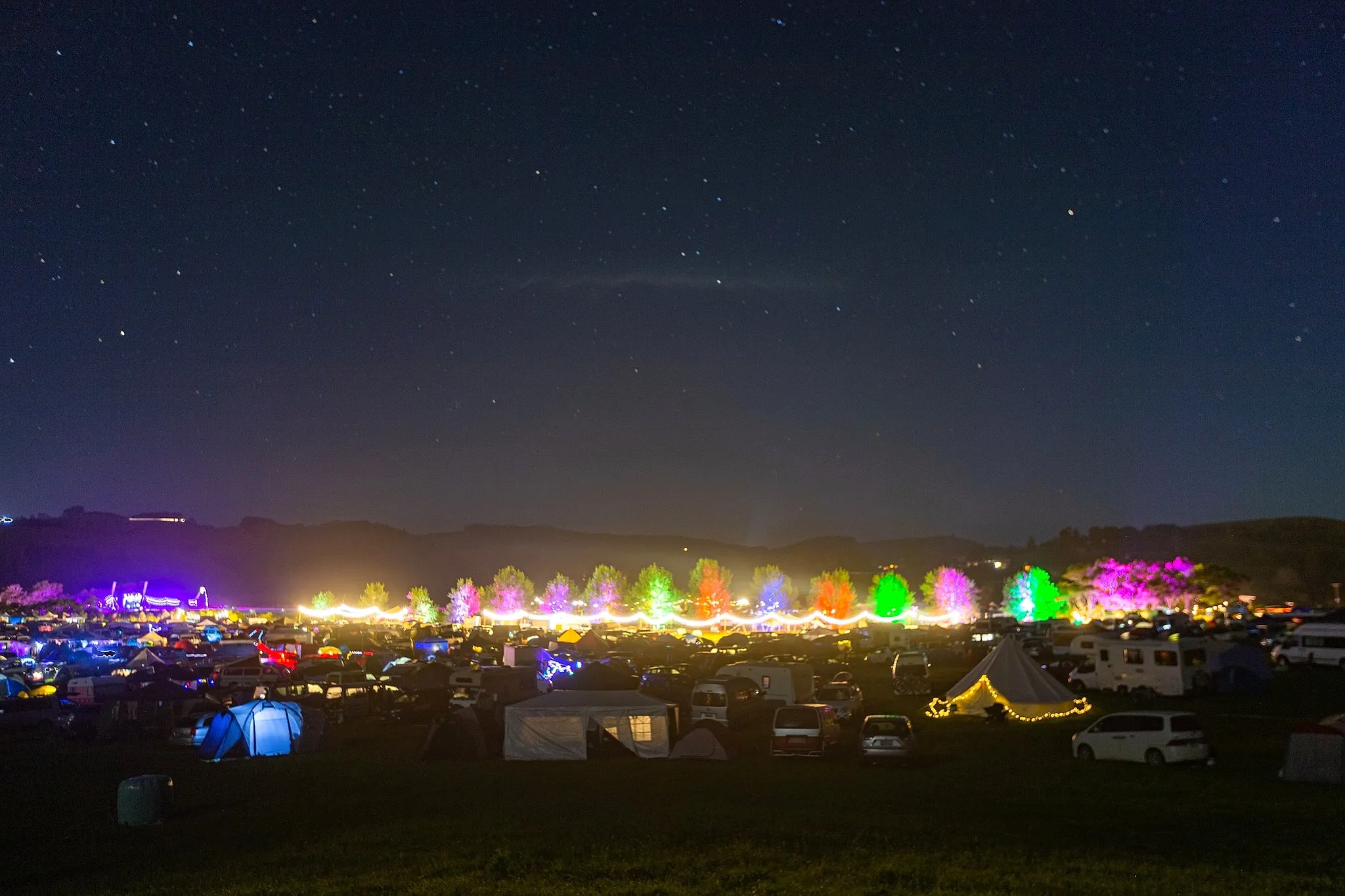 A festival campsite at night beneath a line of colour-washed trees and a warm festoon-lit avenue