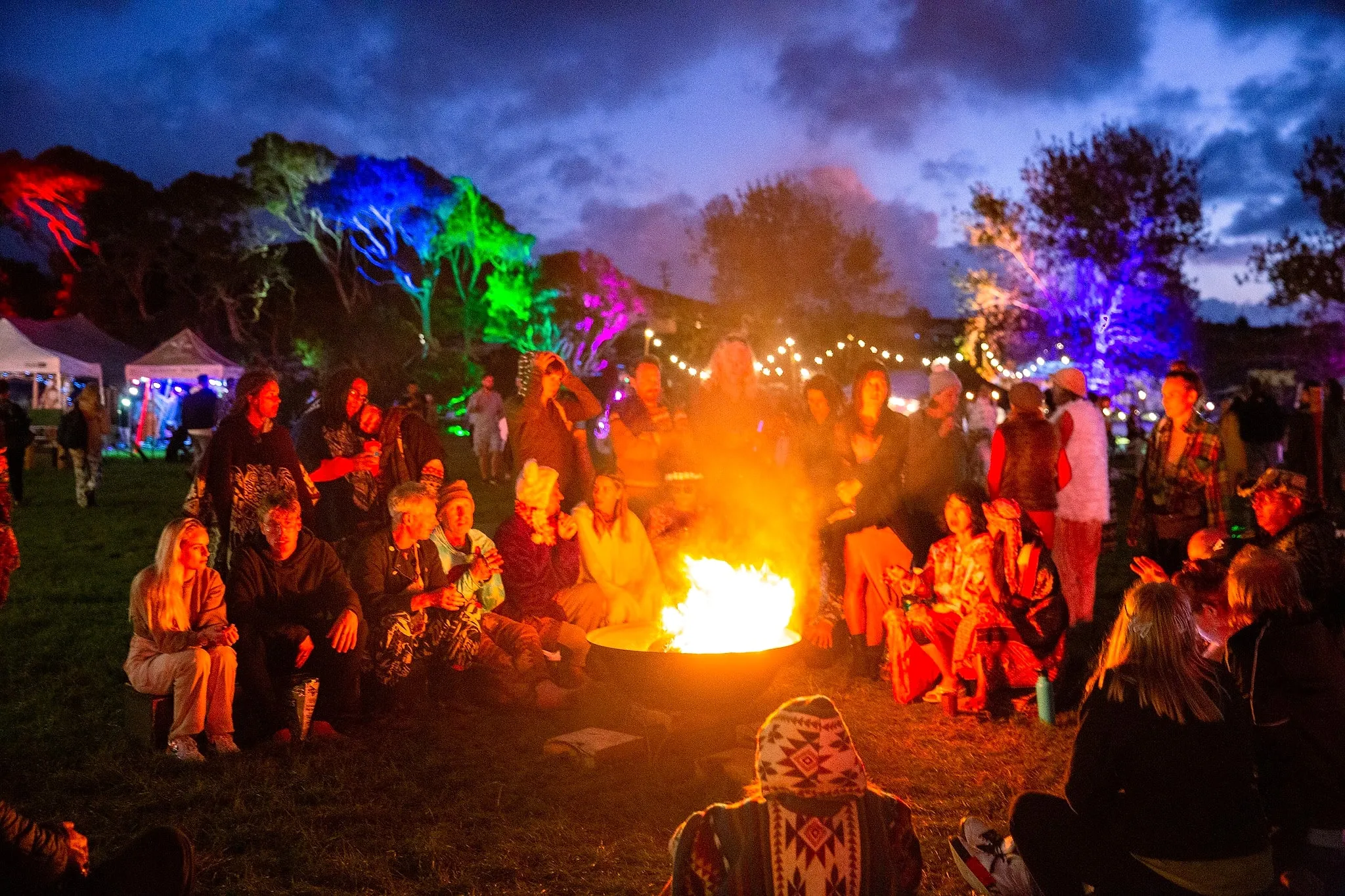 A crowd around a fire bowl at dusk with festoons and colour-washed trees behind